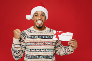 Merry fun young man wear Christmas sweater Santa hat posing hold gift certificate coupon voucher card for store do winner gesture isolated on plain red background. Happy New Year 2023 holiday concept.