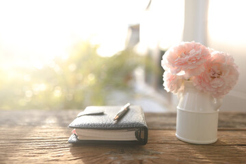 Wall Mural - pink roses in a white vase and leather notebook on wooden table with natural sunlight