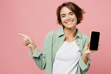 Young happy woman she wear green shirt white t-shirt hold in hand use mobile cell phone with blank screen workspace area point index finger aside isolated on plain pastel light pink background studio.