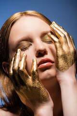 Wall Mural - Portrait of young woman with makeup and golden paint on hands touching face isolated on blue