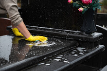 Headstone Cleaning On Cemetery. Professional In Yellow Gloves Cleans The Granite Grave, Scrubbing With Sponge And Water. Tombstone Preparation For All Saints Day On November 1st