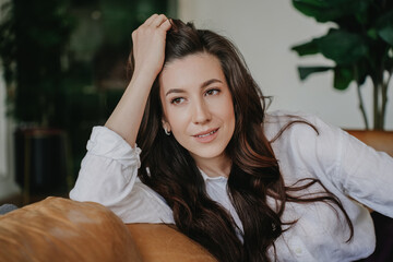 Close up portrait of pretty brunette young adult woman sitting on cozy leather couch looking aside, cute smiling dressed in white shirt. Beautiful hispanic female relaxing at home. Business people.