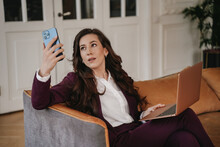 Brunette Businesswoman In White Shirt And Violet Suit Sitting On Cozy Leather Sofa With Laptop Holding Smartphone, Making Video Call. Beautiful Young Entrepreneur Working At Home. Business And Finance
