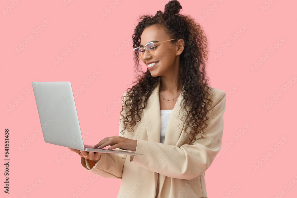 Young African-American woman using laptop on pink background