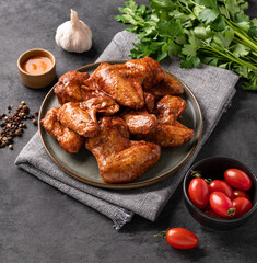 Wall Mural - Spicy grilled chicken wings with sauce on a plate with tomato and herb on a dark background of slate or concrete.
