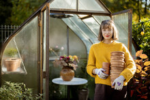 Portrait Of A Young Woman Stands With A Pile Of Clay Jugs In Front Of Glass Orangery At Garden In Sunny Morning. Gardening And Hobby Concept