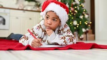 Child Boy In A Santa Hat Lying On A Plaid Smiles Dreamily And Writes A Letter To Santa On A White Sheet - A Wish List On The Background Of A Christmas Tree.