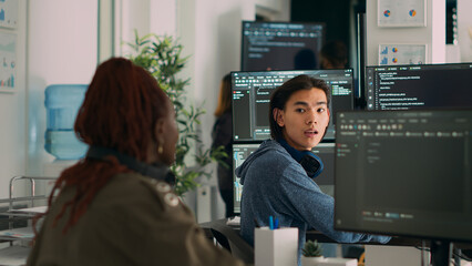 Asian system engineer asking coder for help with html code, pointing at database script on terminal window. Team of diverse developers working on app developing program in data room.