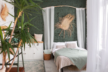 Modern boho interior of bedroom with green brick wall, bed, white wooden commode, curtains, houseplants, rattan basket, dray palm leaf and design personal accessories. Stylish home decor. Template.