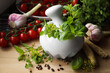 Mortar with fresh herbs near garlic, horseradish roots, black peppercorns and cherry tomatoes on wooden table, closeup