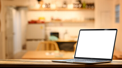 Wall Mural - Laptop on wooden table against blurred kitchen with soft and warm light in background. Blank screen for your advertise design