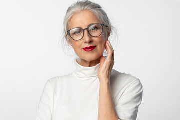 Wall Mural - A lovely elderly intelligent woman with glasses and a slight smile touches her face with her hand. A cosmetic conceptual portrait of aging skin care.