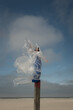 © Milou Dirks - abstract art portrait of woman in long blue dress on beach pole with white plastic sheet, sky and cloud