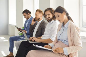 Wall Mural - Portrait of happy man using modern laptop while waiting in line together with other business people. Group of four busy employees sitting in row on office chairs, reading documents or talking on phone