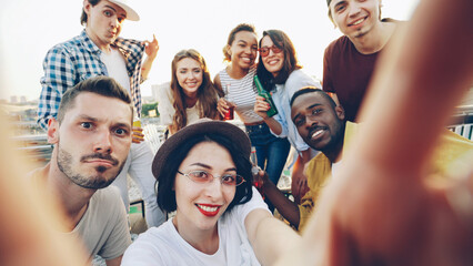 Young woman is taking selfie holding camera and looking at camera while her friends are clinking glasses, posing and showing hand gestures at funny outdoors party.