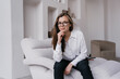 © Iona - Adorable young woman in glasses, white shirt and black pants sitting on cozy chair at home looking at camera toothy smiling. Creative interior designer against interior. Successful people. Business.