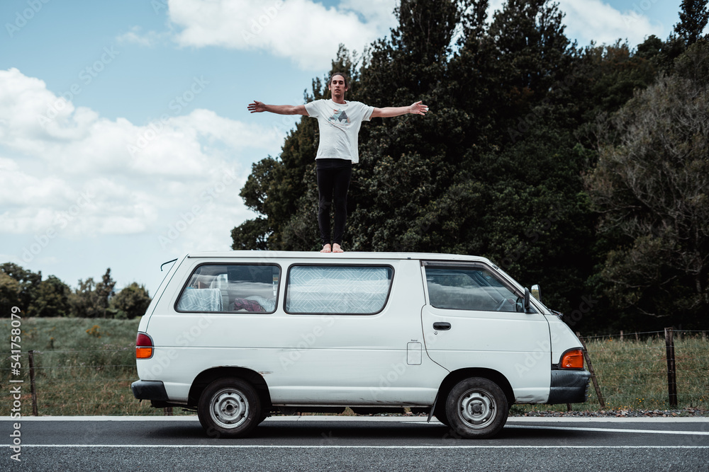 barefoot caucasian young man dressed in a t-shirt and black trousers standing with his arms crossed and feet together on the roof of an old white van, rotorua, new zealand