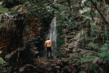 Wall Mural - Caucasian boy with bare back in black pants marveling at the beauty of waterfall water between forest trees on waitawheta tramway, new zealand