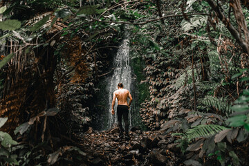 Wall Mural - caucasian young man with long collected hair with bare back and black pants heading to small waterfall with little water between forest trees on waitawheta tramway, new zealand