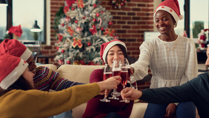 Diverse people clinking alcohol glasses to make toast and celebrate christmas holiday at seasonal office party. Enjoying wine during xmas eve festivity in space with ornaments.