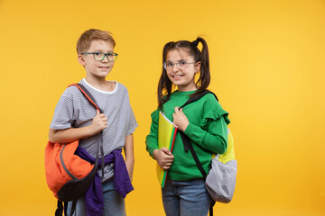 Happy schoolkids wearing trendy outfits posing on yellow background. Smiling kids holding backpacks and books, smiling, looking to camera. Studio shot. Back to school.