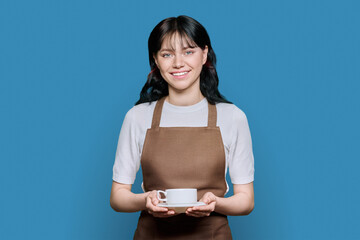 Young smiling female waitress in apron with coffee cup, on blue background