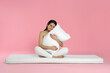 © New Africa - Young woman sitting on soft mattress and holding pillow against pink background