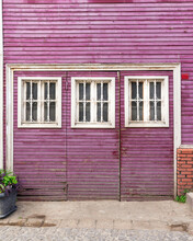 Wooden Grunge Weathered Abandoned Door Painted In Purple, With Three White Wooden Windows Painted In White, In A Cobblestone Alley
