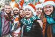 © Davide Angelini - Self portrait of mixed race young people wearing santa claus hat celebrating Christmas day outside - Happy multiracial friends group taking selfie having xmas holiday party - Winter holidays concept