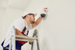 © Studio Romantic - Professional young electrician worker in uniform and cap installs, repairs, screws surveillance video camera on wall in bright office space, school, hospital. He is standing on stepleader, side view.