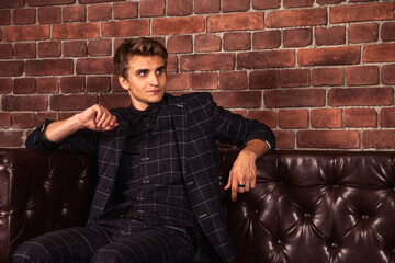 Wall Mural - Caucasian male adult stylish handsome man model in suit indoors. Confident young businessman sitting on sofa looking at camera at brick wall background. Fashion style concept. Copy text space