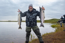 Happy Man Proudly Holds Two Large Salmon Fish Caught On Egegik River In Alaska As Other Fishermen Catch Fish In The Background