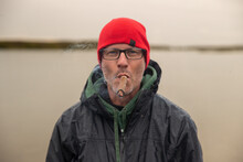 Handsome Man Wearing A Red Beanie Cap Or Hat And Glasses Smokes A Large Cigar Outside Near River In Alaska