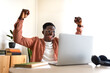 © Daniel - Happy young black male celebrating success with arms raised up in front of laptop.