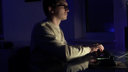 The male programmer works late. A guy with glasses is sitting in a dark office in front of a monitor and writing code.