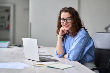 Young happy business woman company employee sitting at desk working on laptop. Smiling female professional entrepreneur worker using computer in corporate modern office looking at camera. Portrait.