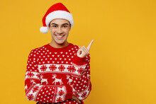 Merry Young Man In Knitted Christmas Sweater Santa Hat Posing Point Index Finger Aside Indicate On Workspace Area Isolated On Plain Yellow Background Happy New Year 2023 Celebration Holiday Concept