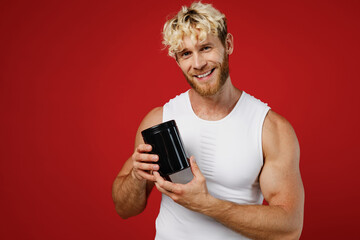 Wall Mural - Young happy strong sporty sportsman toned man wear white clothes spend time in home gym hold black bottle of sport protein supplement isolated on plain red background. Workout sport fit body concept.