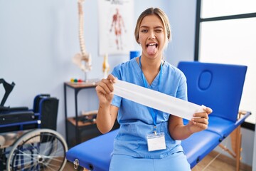 Canvas Print - Young blonde woman holding therapy strip band sticking tongue out happy with funny expression.