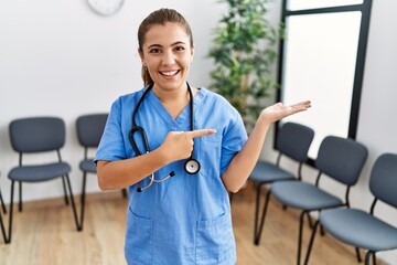 Poster - Young brunette doctor woman at waiting room amazed and smiling to the camera while presenting with hand and pointing with finger.