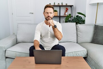 Poster - Middle age man using laptop at home with hand on chin thinking about question, pensive expression. smiling with thoughtful face. doubt concept.