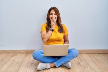 Sticker - Hispanic woman using laptop sitting on the floor at home laughing and embarrassed giggle covering mouth with hands, gossip and scandal concept