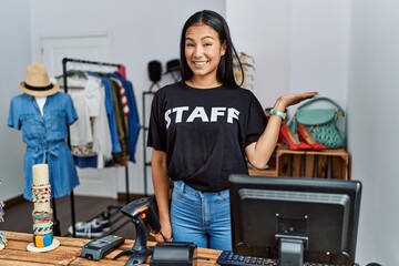 Sticker - Young hispanic woman working as staff at retail boutique smiling cheerful presenting and pointing with palm of hand looking at the camera.