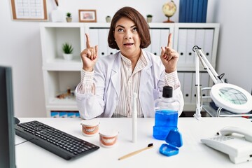 Canvas Print - Middle age hispanic dentist woman working at medical clinic amazed and surprised looking up and pointing with fingers and raised arms.