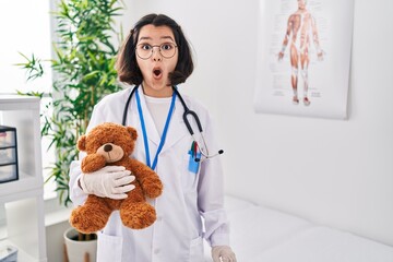 Canvas Print - Young hispanic doctor woman holding teddy bear scared and amazed with open mouth for surprise, disbelief face