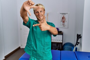 Poster - Young blond man wearing physiotherapist uniform standing at clinic smiling making frame with hands and fingers with happy face. creativity and photography concept.