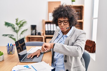 Wall Mural - Black woman with curly hair wearing call center agent headset at the office checking the time on wrist watch, relaxed and confident