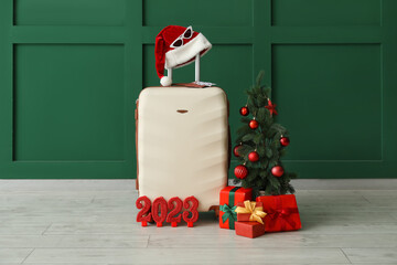 Suitcase with Santa hat, Christmas tree, gifts and figure 2023 near green wall