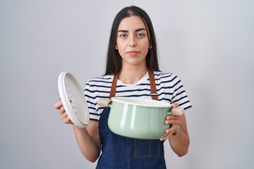 Sticker - Young brunette woman wearing apron holding cooking pot relaxed with serious expression on face. simple and natural looking at the camera.