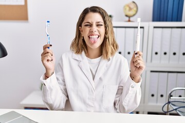 Canvas Print - Young hispanic dentist woman holding electric toothbrush and teethbrush at clinic sticking tongue out happy with funny expression.